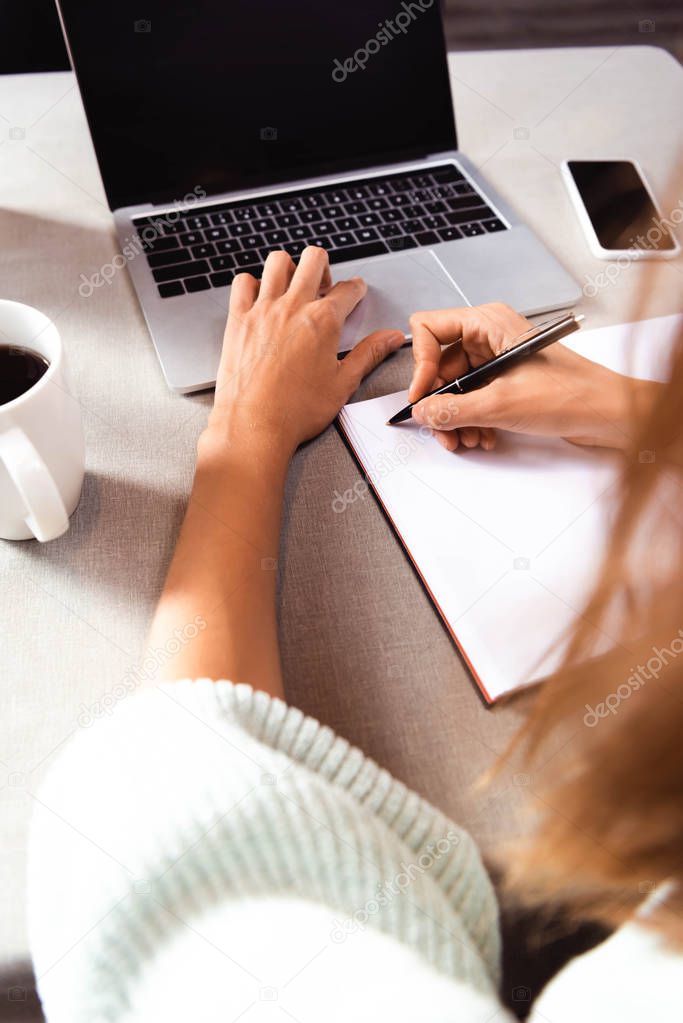 cropped view of woman writing in notepad in cafe with coffee cup, smartphone and laptop