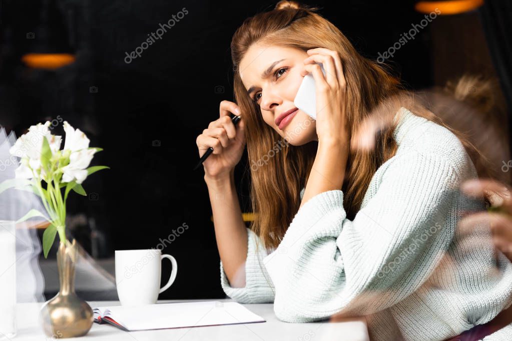 thoughtful woman talking on smartphone in cafe with notepad and cup of coffee