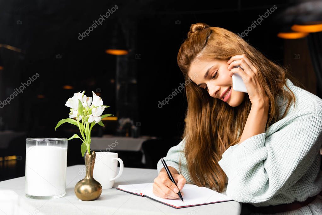 smiling girl talking on smartphone and writing in notepad in cafe and coffee cup