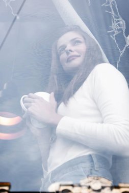 Pencerenin arkasındaki kafede bir fincan kahve tutan genç bir kadının seçici odak noktası.