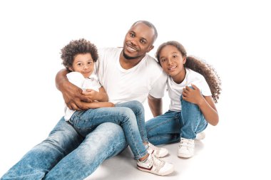 happy african american man embracing son and daughter while sitting together on white background clipart