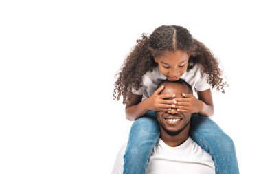 Neşeli Afrikalı Amerikalı çocuk babasının gözlerini elleriyle kapatırken beyazı sırtında taşıyor.