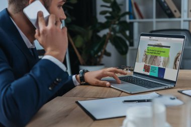 Kyiv, Ukrayna - 1 Ekim 2019: Dizüstü bilgisayarın yanında oturan genç işadamı Bbc Science web sitesi ekranda ve akıllı telefondan konuşuyor.
