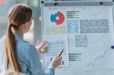 back view of young businesswoman pointing with pen at infographics on flipchart