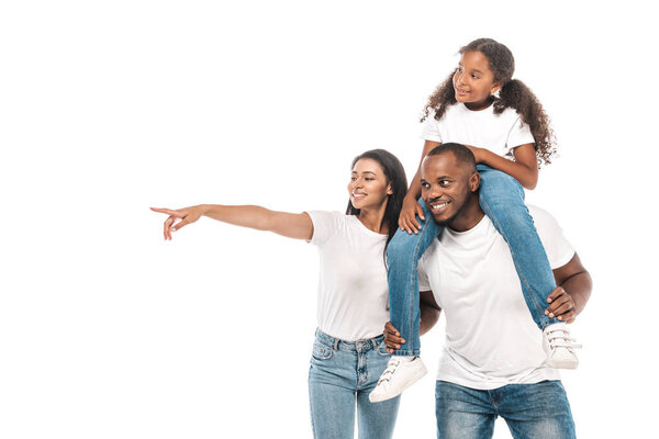 cheerful african american woman pointing with finger, and husband piggybacking adorable daughter isolated on white