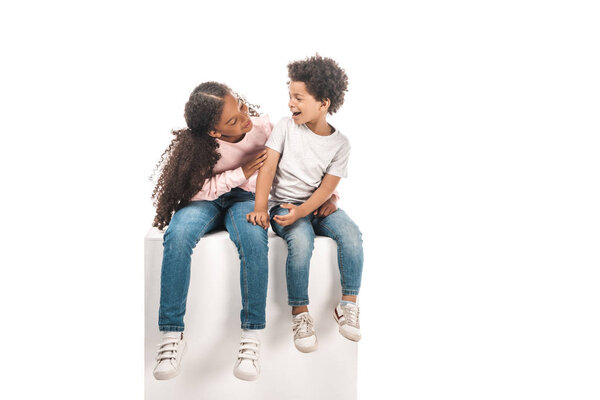 cute african american sister looking at adorable brother while sitting on white cube together isolated on white