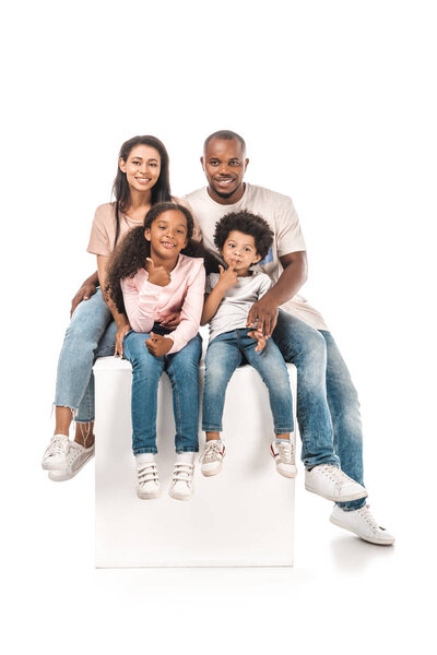 cheerful african american child showing thumb up while sitting on white cube with parents and brother on white background