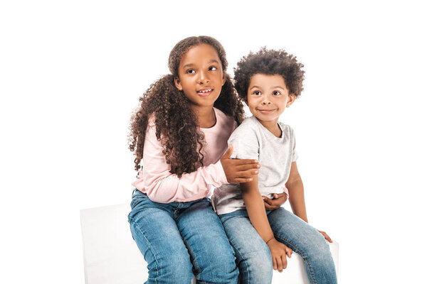 cute african american sister hugging adorable brother while sitting on pedestal and looking away together isolated on white