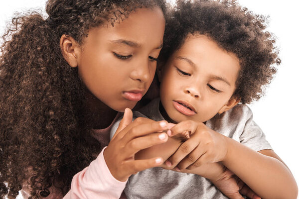 attentive african american sister looking at hand of adorable brother isolated on white