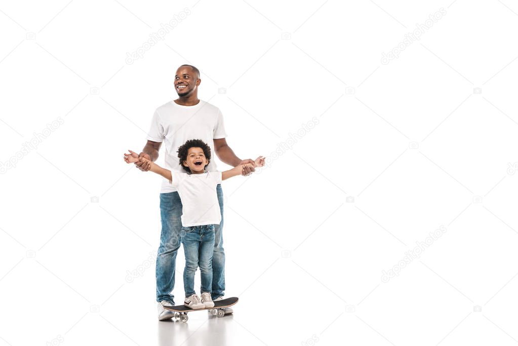 happy african american man supporting cheerful son standing on penny board on white background