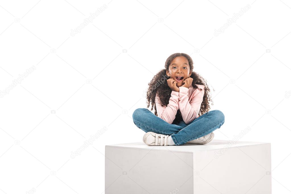 cheerful african american child sitting on white cube with crossed legs isolated on white