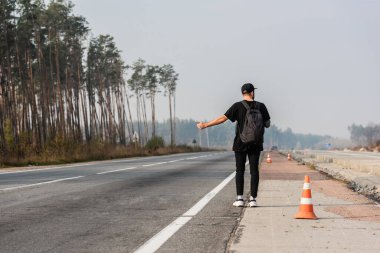 Yeşil ağaçların yanında otostop çekerken başparmağını gösteren şapkalı bir adamın arka görüntüsü 