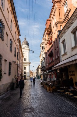 LVIV, UKRAINE - OCTOBER 23, 2019: people walking along narrow street and carmelite church against blue sky clipart