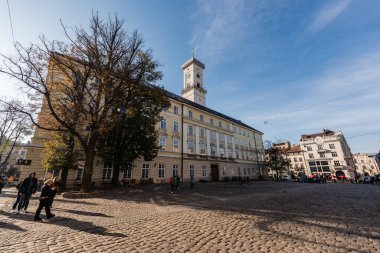 LVIV, UKRAINE - OCTOBER 23, 2019: facade of lviv city hall tower and people walking along street clipart