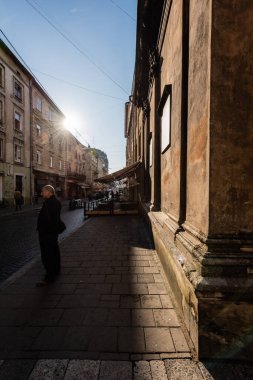 LVIV, UKRAINE - 23 Ekim 2019: Parlak güneşli mavi gökyüzüne karşı sokakta ve eski evlerde duran adam