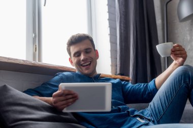 Neşeli bir adam elinde kahve fincanıyla dijital tablete bakıyor.