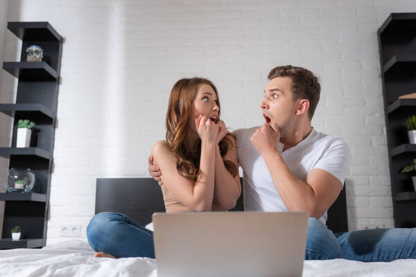 scared couple looking at each other near laptop in bedroom 