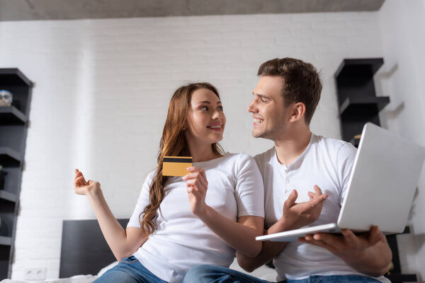 happy girl holding credit card and man using laptop while looking at each other 