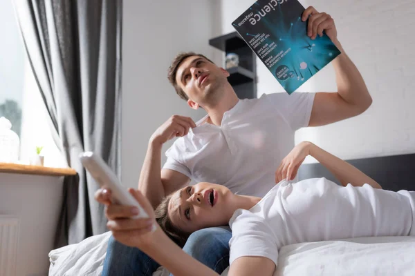 エアコンのリモコンを持った若い女性が熱い感じながら雑誌を持ってベッドの上に横になって — ストック写真