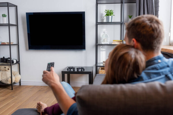 Избирательный взгляд мужчины, держащего пульт дистанционного управления и смотрящего на плоский панельный телевизор с бланкетным экраном рядом с женщиной в гостиной
  
