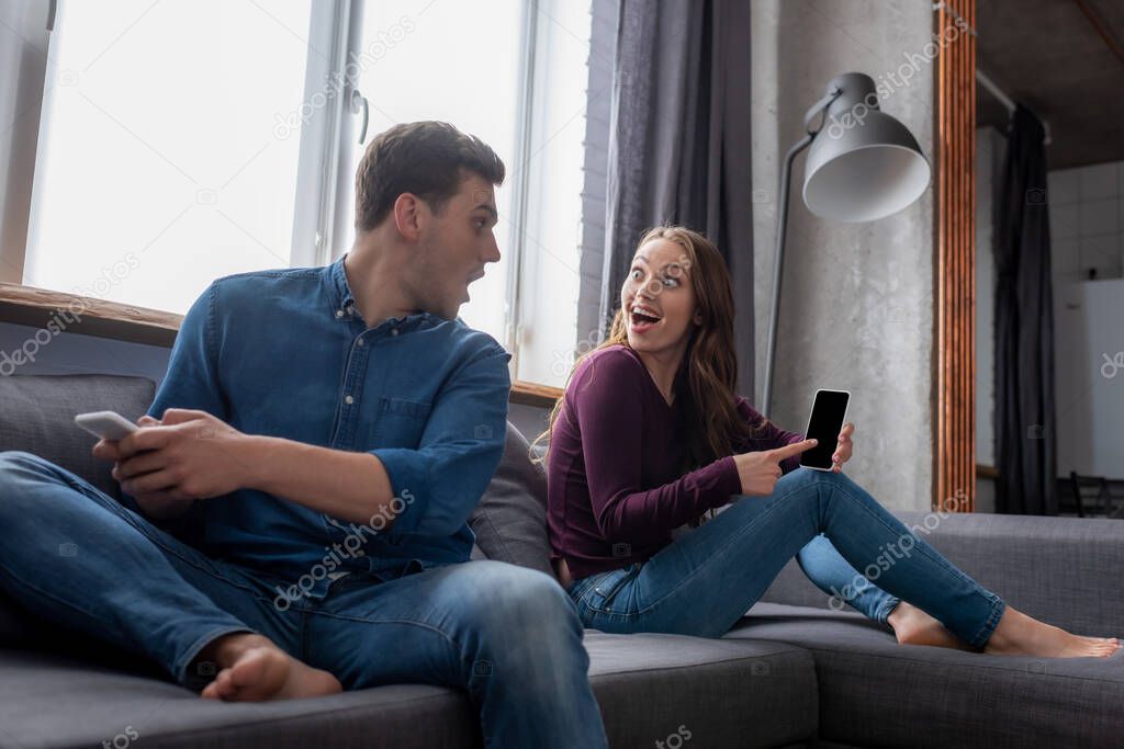 excited girl pointing with finger at smartphone with blank screen and looking at boyfriend in living room 