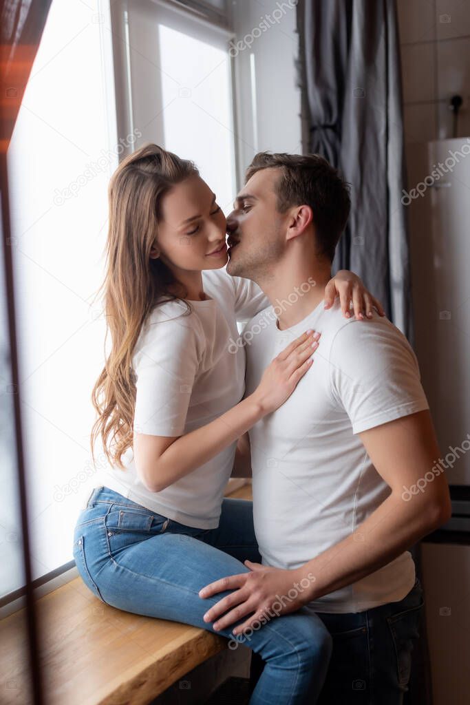selective focus of handsome man kissing young woman sitting on window sill at home