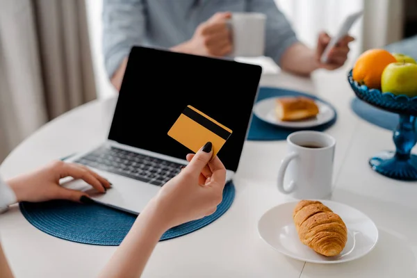 女孩在笔记本电脑旁拿着信用卡 桌上有空白屏幕和早餐的剪影 — 图库照片
