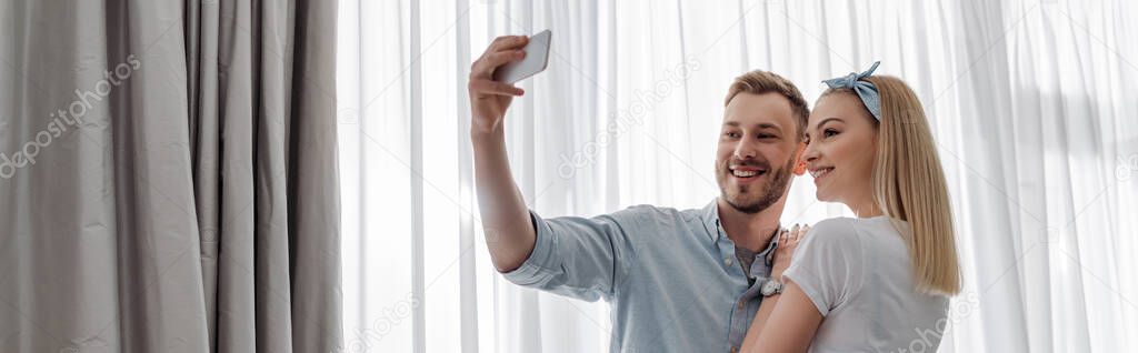 panoramic crop of couple smiling while taking selfie at home 