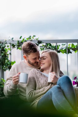 selective focus of happy man and smiling woman holding cups of tea and sitting on outdoor sofa  clipart