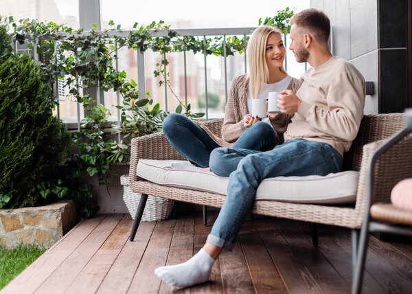 счастливые женщина и мужчина смотрят друг на друга и держат чашки, сидя на открытом диване
