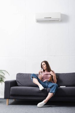 Yorgun bir kadın, evinde klimayla sıcaktan acı çekerken su içiyor. 