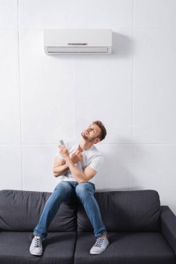 confused man holding remote controller and suffering from heat with broken air conditioner at home  clipart