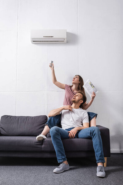 exhausted couple with remote controller and newspaper suffering from heat while sitting home with broken air conditioner 