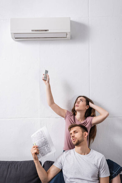 young couple with remote controller and newspaper suffering from heat while sitting home with broken air conditioner 