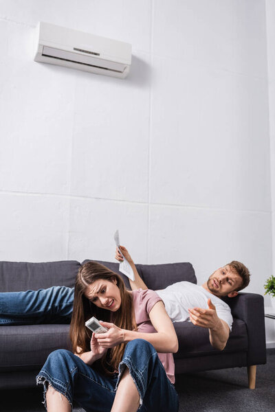 young couple with remote controller and newspaper suffering from heat while sitting home with broken air conditioner 