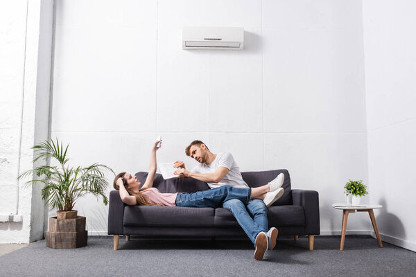 sad young couple with remote controller suffering from heat at home with broken air conditioner 