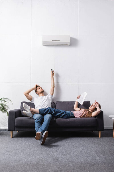 young tired couple with remote controller suffering from heat at home with broken air conditioner 