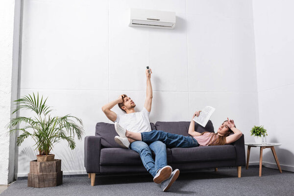 frustrated young couple with remote controller suffering from heat at home with broken air conditioner 