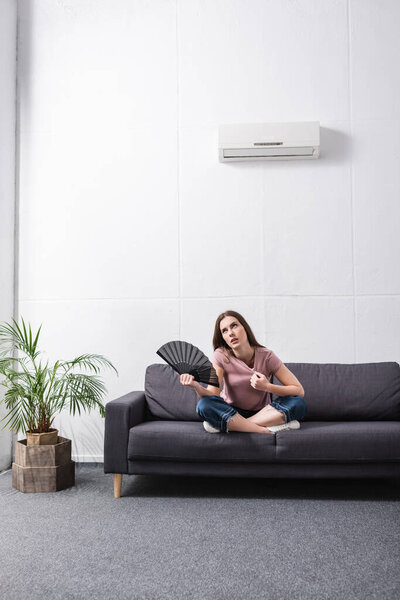 upset young woman suffering from heat and using hand fan at home with broken conditioner