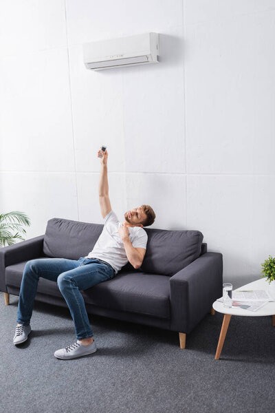 sad man holding remote controller and suffering from heat with broken air conditioner at home 