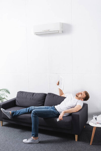 exhausted man suffering from heat while using newspaper as hand fan at home with broken air conditioner
