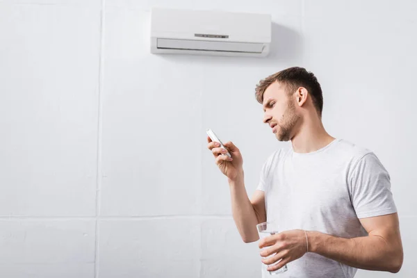 Uomo Frustrato Che Tiene Bicchiere Acqua Mentre Cerca Accendere Condizionatore — Foto Stock