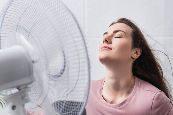 attractive positive girl feeling comfortable with electric fan at home during summer heat 