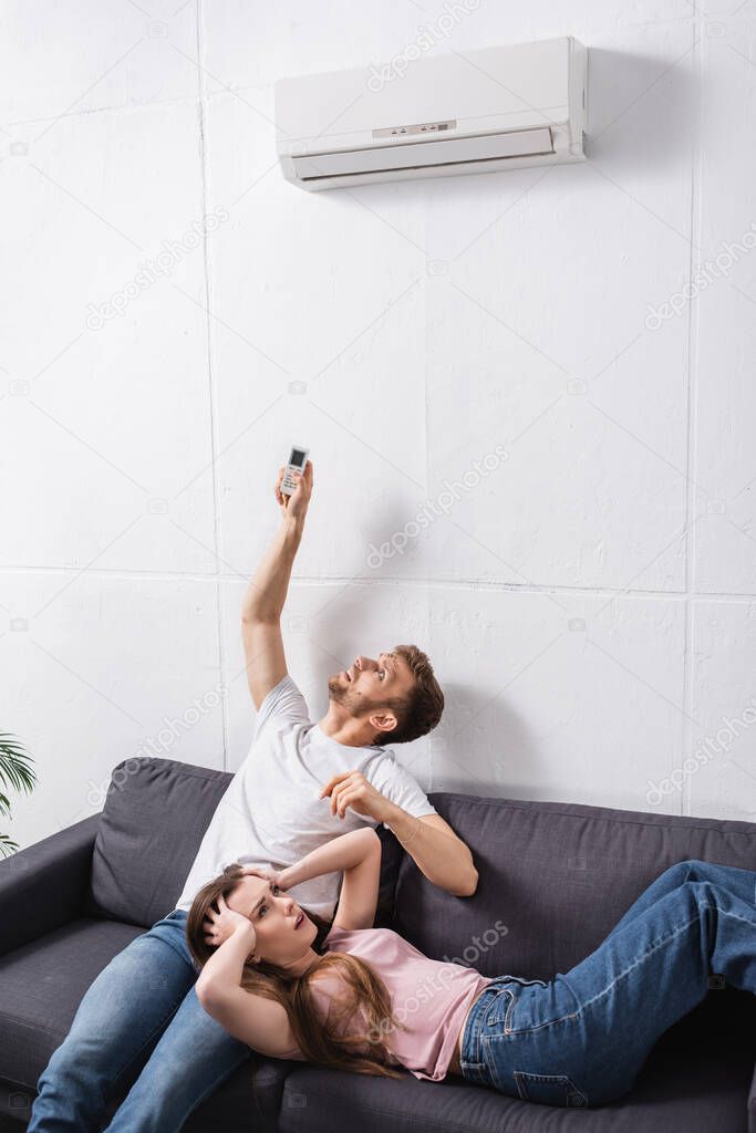 Frustrated young couple with remote controller suffering from heat at home with broken air conditioner