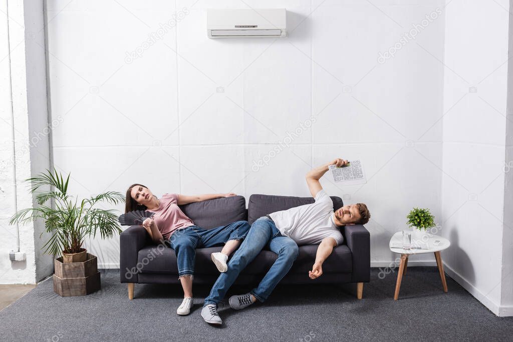 sad couple with hand fan and newspaper suffering from heat at home with broken air conditioner 