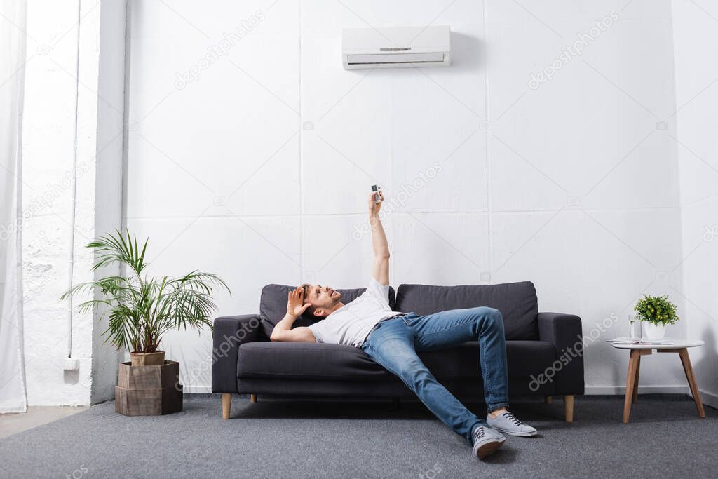 exhausted man holding remote controller and suffering from heat with broken air conditioner at home 