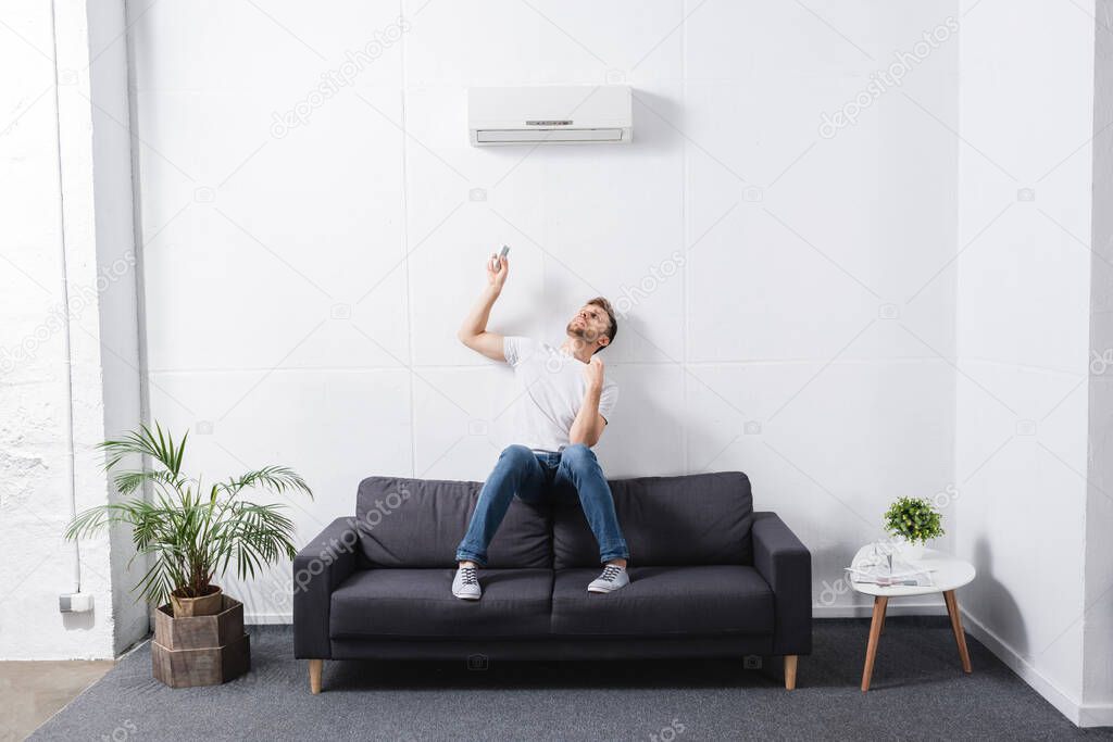 tired man holding remote controller and suffering from heat with broken air conditioner at home 