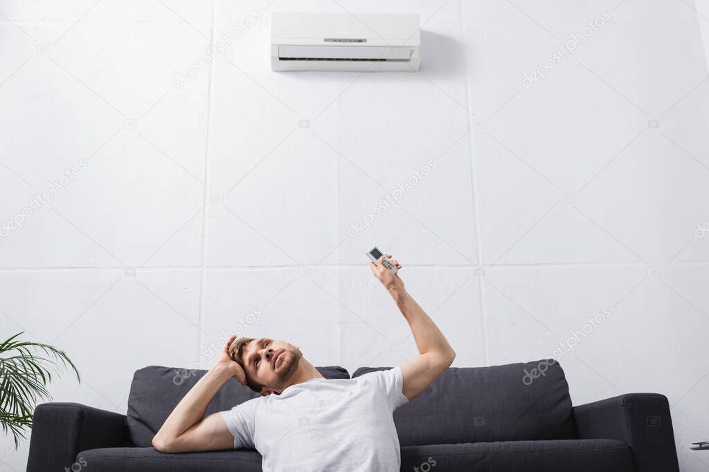 frustrated man holding remote controller and suffering from heat with broken air conditioner at home 