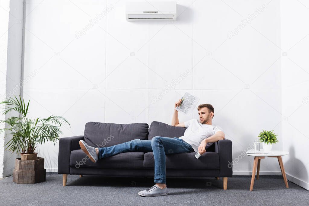 young man suffering from heat while using newspaper as hand fan at home with broken air conditioner