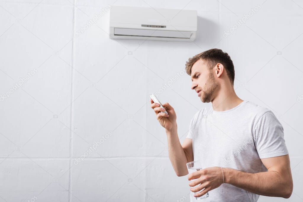 frustrated man holding glass of water while trying to switch on air conditioner with broken remote controller at home 
